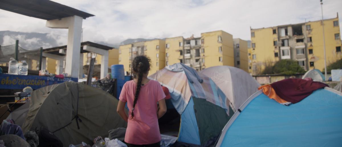 Terremotos en Venezuela 2026: Una niña camina entre tiendas de campaña en un asentamiento improvisado con edificios derruidos al fondo.